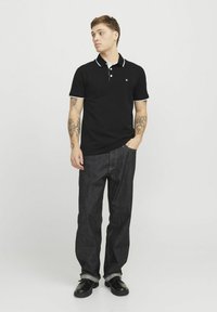 Zwarte poloshirt met witte kraag en accenten op de mouwen, gecombineerd met donker denim jeans. Model staat iets opzij gericht.