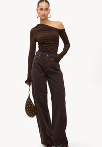 Haut marron asymétrique à une épaule avec une texture plissée, associé à un jean large taille haute marron foncé. Accessoires comprenant un sac marron avec des accents dorés.
