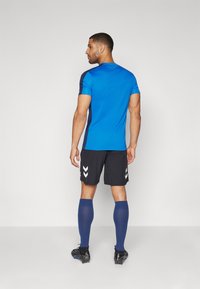 Sportos öltözék, amely egy kék rövid ujjú pólóból, fekete rövidnadrágból fehér ék részletekkel, és kék térdig érő zoknikból áll. Fekete focicipők.