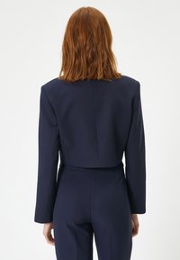 Marineblå cropped blazer med lange ærmer, skræddersyet design, tekstureret stof og subtile sømme på ryggen, stylet over et matchende outfit.