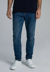 Niebieskie dżinsy o slim fit, z przeszywanymi akcentami, w tradycyjnym pięciokieszeniowym wzorze, noszone z białymi trampkami.