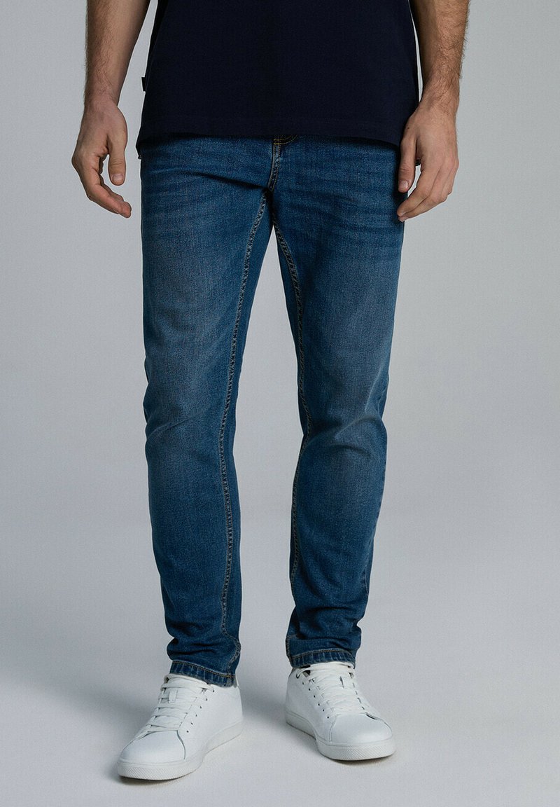 Niebieskie dżinsy o slim fit, z przeszywanymi akcentami, w tradycyjnym pięciokieszeniowym wzorze, noszone z białymi trampkami.