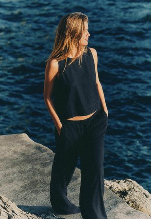 Tânără în top negru fără mâneci și pantaloni, stând pe o țărm stâncos, privind spre dreapta, cu valuri oceanice pe fundal.