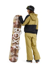 Snowboard con un design scenico della foresta e il bold "ROXY" in bianco. Il modello indossa una giacca con cappuccio bicolore e pantaloni da neve gialli.