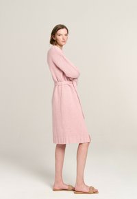 Helles rosa Strickgewand mit langen Ärmeln, tailliertem Gürtel und geripptem Saum. Das Model trägt helle braune Slipper. Weicher Stoff.