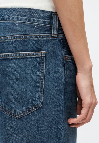 Mâna stângă odihnindu-se lângă o pereche de blugi albaștri din denim, cu buzunarul din spate și bucla pentru curea vizibile, purtând un top alb cu reieșiri tip rib.