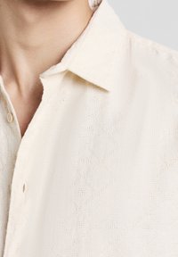 Primo piano di una persona che indossa una camicia button-up di colore crema con una texture, con colletto a punta, che mostra il collo e la parte superiore del torace.