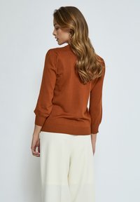 Pull en tricot marron avec des manches trois-quarts et des poignets côtelés. Présente une texture douce, une coupe ajustée et un col rond.