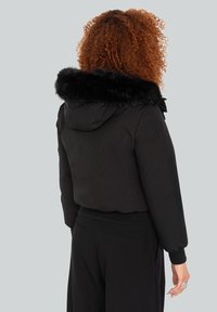 Sort cropped jakke med en faux fur hætte og manchetter. Har et tætsiddende design, glat tekstur og fremtrædende syninger langs sømmene.