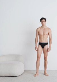 Caleçons noirs avec une ceinture logo, le modèle a une silhouette tonique, se tient sur un tapis clair à côté d'un canapé incurvé et doux de couleur blanche.