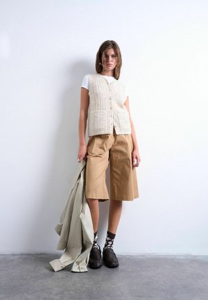 Jonge vrouw staat tegen een witte muur, draagt een beige gebuttonneerde vest, wit overhemd, tan culottes, zwarte schoenen met patroon sokken, houdt een jas vast.