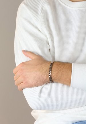 Silver braided bracelet on a wrist, featuring a textured design with polished accents, worn over a white textured long-sleeve shirt.