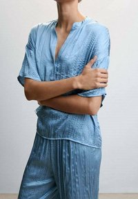 Top azzurro chiaro a maniche corte con scollatura a V, realizzato in tessuto testurizzato. Il top presenta pieghe sottili e un taglio rilassato.