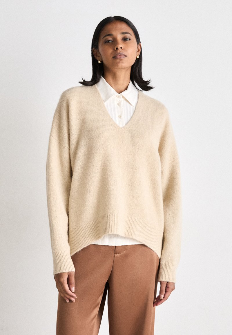 Beige oversized sweater with a V-neckline, ribbed hem, and long sleeves, paired with a white collared shirt underneath and brown trousers.