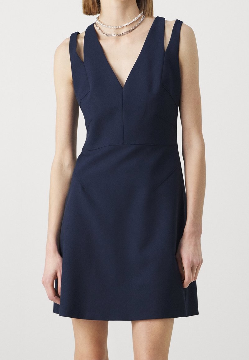 Femme portant une robe bleu marine sans manches avec un profond décolleté en V et des bretelles asymétriques, associée à des colliers superposés de perles et de chaînes.
