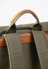 Borsa verde oliva con superficie texturizzata, dettagli in pelle sul manico e etichetta con logo impresso. Maniglie robuste e zip liscia.