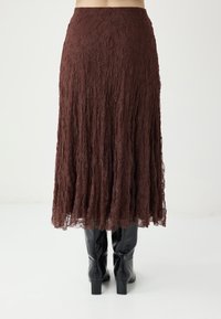 Jupe en dentelle marron avec un design structuré et fluide. La jupe tombe sous le genou, associée à des bottes noires mi-hautes à talons blocs.