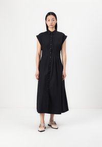 Vestido midi preto, com botões e mangas curtas, apresentando um corpete ajustado e uma saia evasê. Usado com sapatos de cor clara.