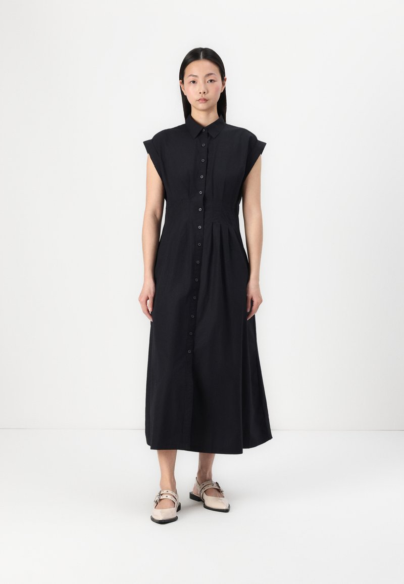 Vestido midi preto, com botões e mangas curtas, apresentando um corpete ajustado e uma saia evasê. Usado com sapatos de cor clara.