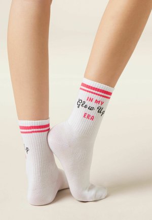 Benedenbenen met witte enkelsokken met roze strepen en de tekst "IN MY Glow Up ERA," staand op een lichtgekleurde vloer tegen een effen achtergrond.