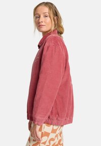Giacca in velluto a coste rosa con colletto, presenta una texture a coste e polsini elasticizzati. Il modello indossa pantaloni a righe crema e arancione.