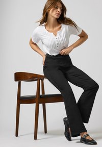 Top blanco de manga corta con tapeta y pliegues, combinado con pantalones negros de talle alto. Modelo sentado en una silla de madera con cojín negro.
