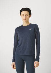 Sötétkék, hosszú ujjú sportpóló, amely struktúrált anyagból készült, és egy kis fényvisszaverő logóval van ellátva. Sötétkék, testhezálló leggingssel párosítva.