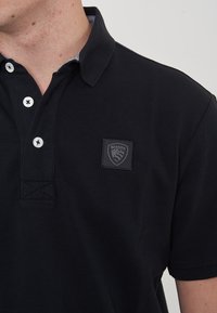 Polo nero con tessuto lavorato, chiusura a bottone e una patch con logo a scudo sul petto. Il colletto presenta un design interno a contrasto.