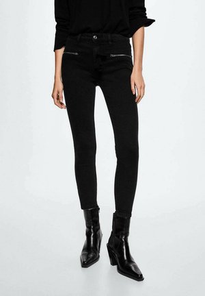 Persoon draagt zwarte skinny jeans met ritszakken aan de voorkant en zwarte enkellaarsjes met hak, staand tegen een effen achtergrond.