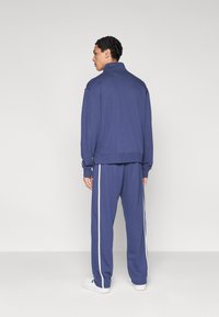 Σκούρο μπλε φόρμα με υψηλό γιακά και λευκές ρίγες στα πλαϊνά; διαθέτει άνετη εφαρμογή και ριμπ μανσέτες τόσο στο μπουφάν όσο και στο παντελόνι.