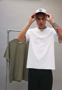 Jeune homme aux bras tatoués portant un t-shirt blanc et une casquette, ajustant sa casquette, avec un t-shirt vert olive accroché à un portant métallique derrière lui.