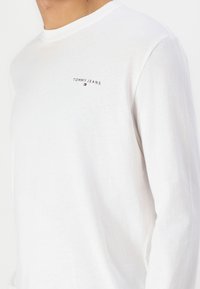 T-shirt à manches longues blanche en coton doux, avec un logo "TOMMY JEANS" brodé en noir au-dessus d'un petit emblème rouge et bleu.