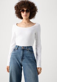 Camisa blanca de manga larga con un escote amplio, combinada con jeans de mezclilla azul de cintura alta. La modelo lleva unas gafas de sol hexagonales grandes.