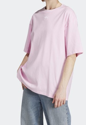 Osoba nosząca luźną, jasnoróżową koszulkę Adidas z małym logo oraz jasnoniebieskie jeansy, stojąca z jedną ręką w kieszeni.