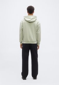 Άνδρας που στέκεται με την πλάτη γυρισμένη, φοράει ανοιχτό πράσινο φούτερ με κουκούλα, μαύρο παντελόνι και σκούρα παπούτσια, μπροστά σε απλό λευκό φόντο.