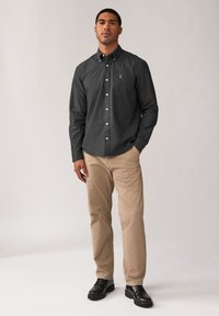 Camicia a maniche lunghe grigio scuro con bottoni e un logo sottile, abbinata a pantaloni beige chiaro e scarpe nere, in piedi di fronte a uno sfondo neutro.