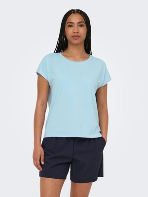 Vrouw draagt een lichtblauw T-shirt met korte mouwen en marineblauwe broekjes, staand met één hand in de zak tegen een effen witte achtergrond.