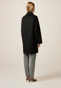 Cappotto di lana nero con colletto, vestibilità ampia e spalle basse, abbinato a pantaloni a quadretti e scarpe con tacco nere.