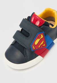 Sötétkék bőr sportcipő sárga belsővel, piros részletekkel és Superman logóval. Téptetős pántokkal és gumis orrmerevítővel rendelkezik.