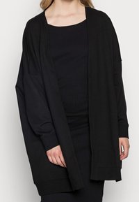 Woman wearing a loose black open-front cardigan over a fitted ribbed black dress against a plain background.