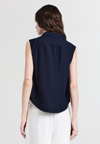 Donna con lunghi capelli castani che indossa una blusa senza maniche blu navy e pantaloni bianchi, vista da dietro su uno sfondo chiaro e uniforme.