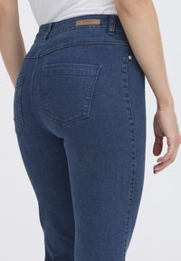 Jean en denim bleu foncé avec taille haute, poches arrière et un patch logo sur la taille. Le tissu a une texture lisse.
