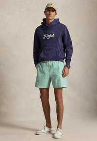 Noor mees, kes kannab tumesinist Ralphi kapuutsi, rohelist ruudulistes lühikutes, beežist nokamütsi, valgeid tossusid ja käekella, seisab neutraalsel taustal.