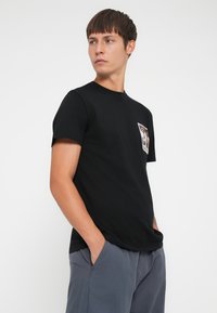 Black cotton t-shirt with a crew neckline, short sleeves, and a colorful graphic patch on the chest. Worn with light gray pants.