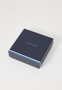 Boîte cadeau carrée bleu marine avec une texture lisse ; présente un bandeau rayé blanc, rouge et bleu le long du bord et le logo "TOMMY HILFIGER" sur le dessus.