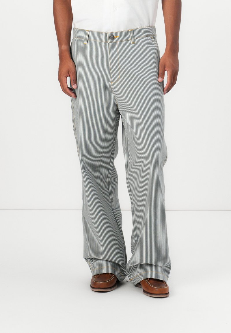 Striped pants in light blue and white, featuring a relaxed fit, button closure, and sturdy fabric with pockets on the sides. Brown shoes worn.