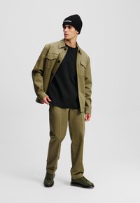 Completo verde oliva composto da una camicia con bottoni e due tasche sul petto, una camicia nera con texture, pantaloni coordinati e scarpe verde scuro.