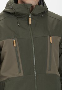 Olivgrön vattentät jacka med dragkedja framtill, två bröstfickor med dragkedjestängning och justerbar huva. Texturerade tygdetaljer.