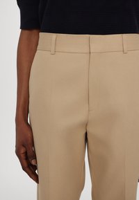 Bezs színű, slim fit szabású nadrág, lapos elülső részű, oldalsó zsebekkel és övhurkokkal, sima anyagból, egy kéz tartja.