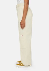 Beige wijde broek gemaakt van duurzaam materiaal, met een zijzak en logo patch, gecombineerd met gele sneakers op een witte achtergrond.